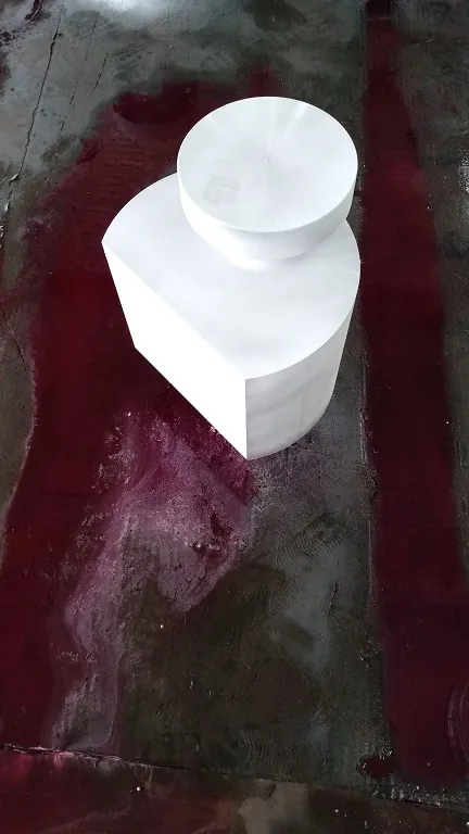 A machined block forging with a stepped round boss, fully coated in white developer, standing on a floor stained red