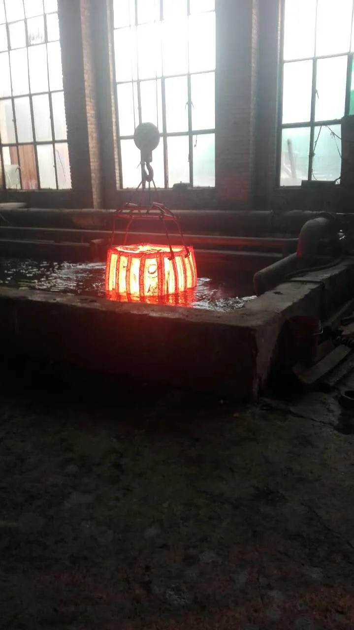A glowing load of parts in a steel lifting frame hanging from a crane hook, being lowered into a liquid-filled quench tank