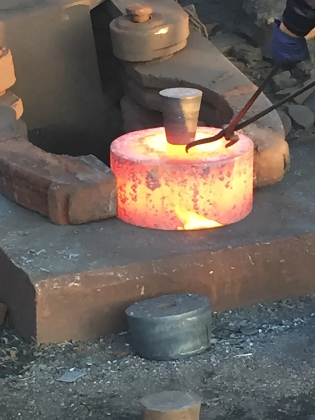 A glowing cylindrical forging blank on the anvil with a tapered punch standing in its top; a gloved hand holds tongs
