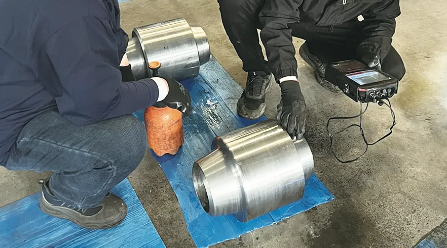 Two inspectors crouch beside machined stepped cylindrical forgings; one presses an ultrasonic probe onto a forging