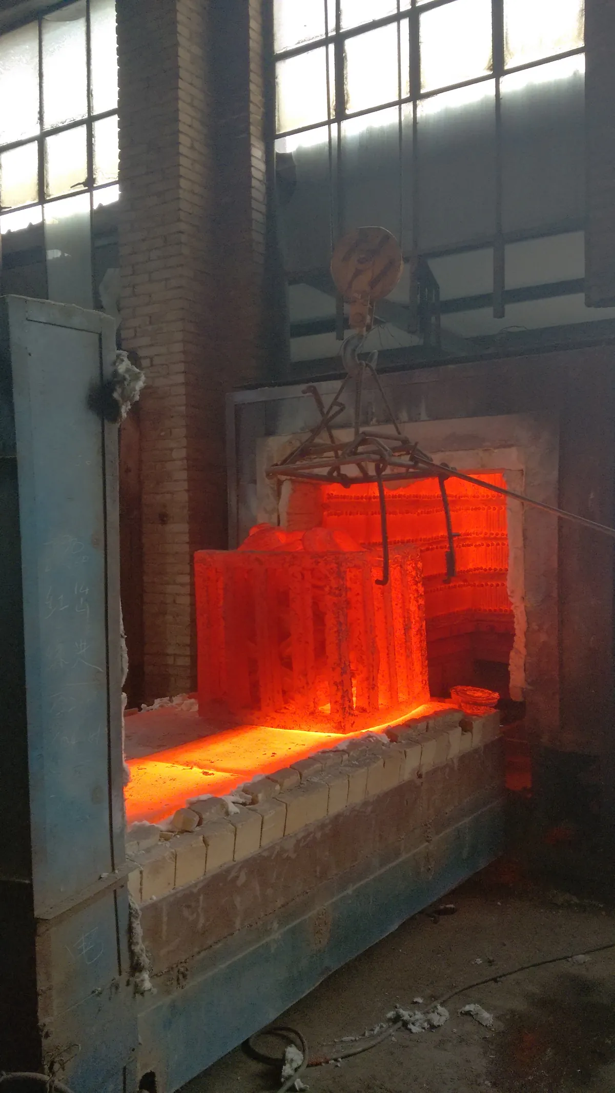 A glowing load of parts in a steel lifting frame being raised by crane from an open car-bottom furnace