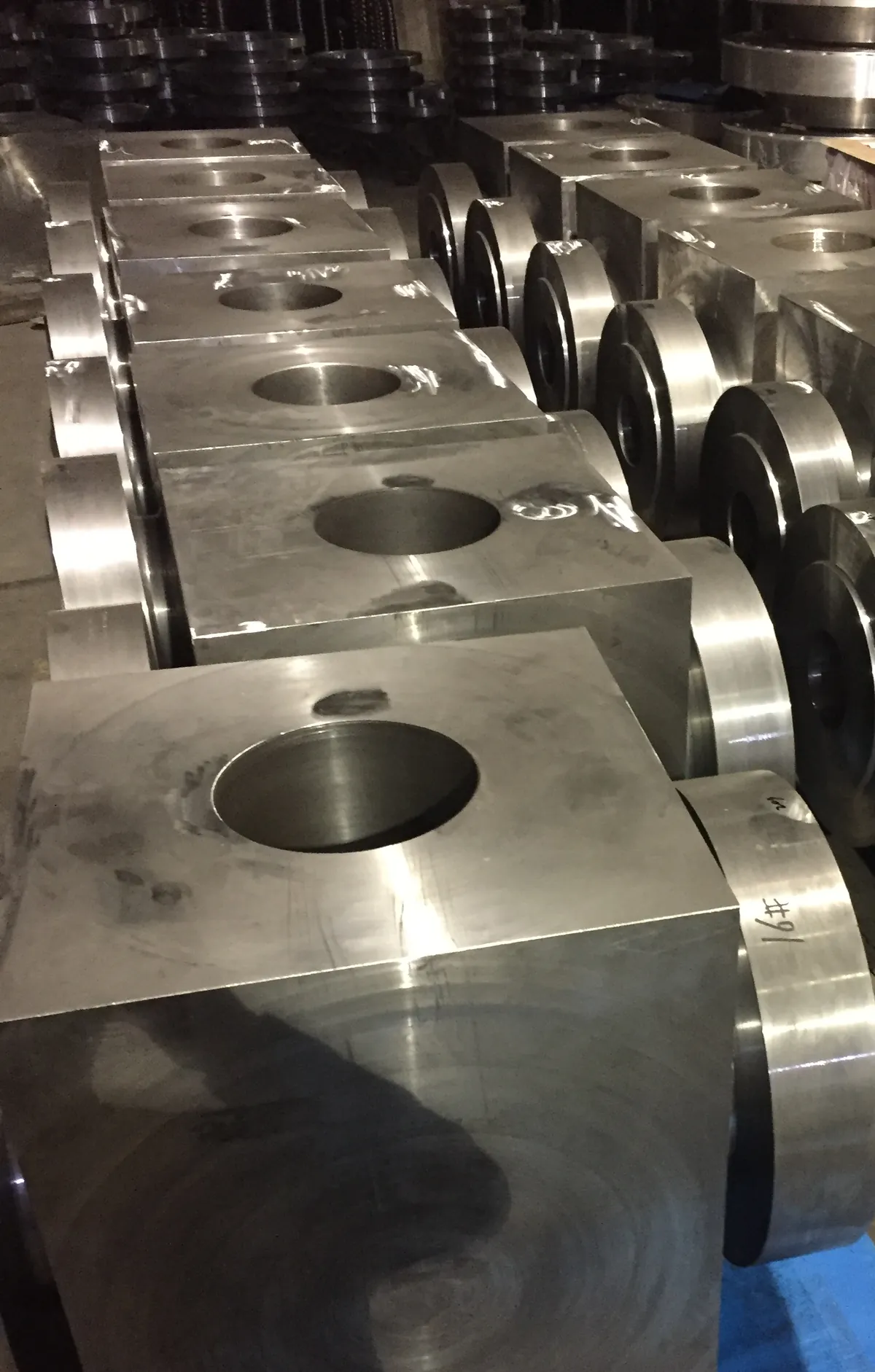 Rows of machined rectangular block forgings with bored holes, and machined cylindrical parts, on a workshop floor