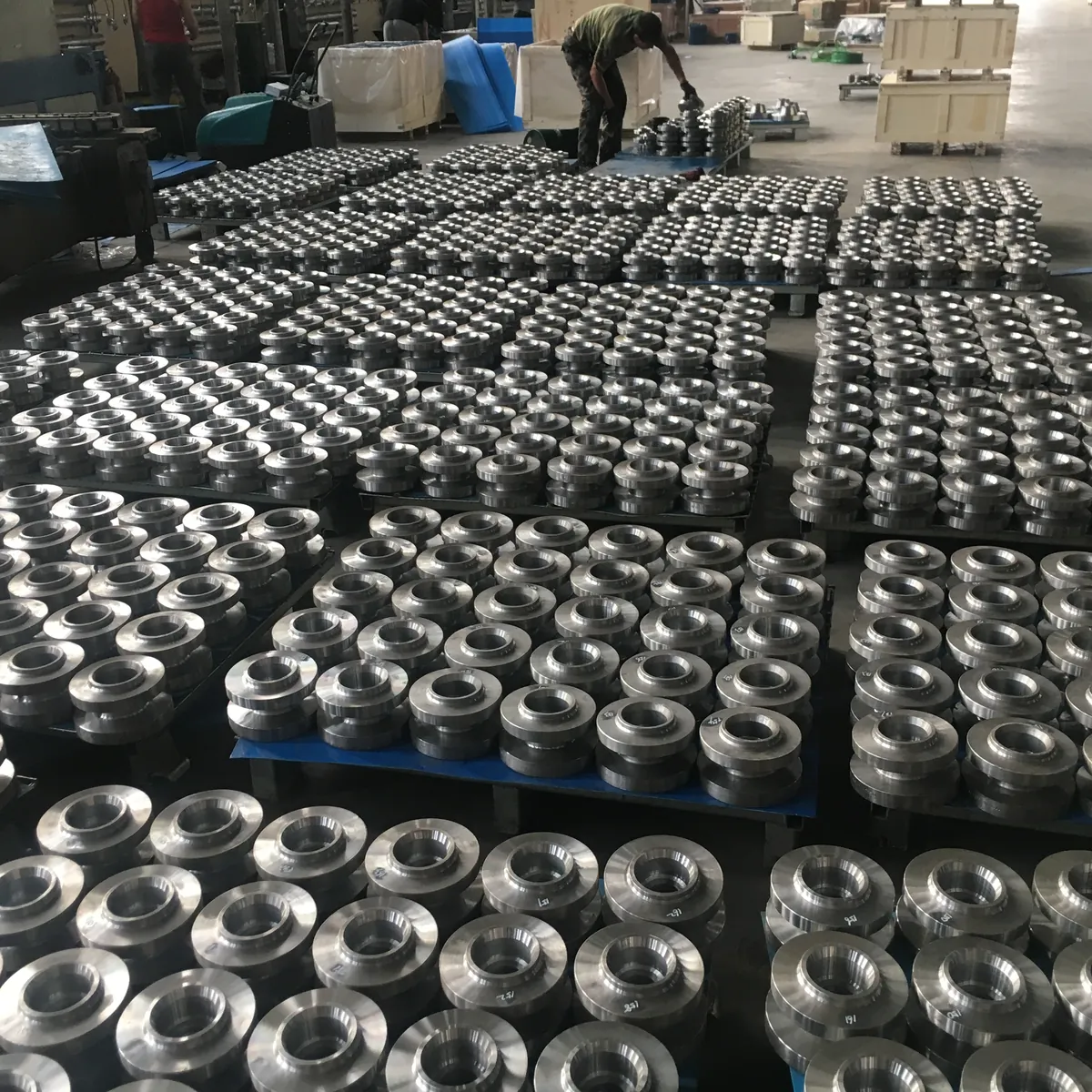Hundreds of machined weld neck flanges laid out on pallets on a workshop floor, wooden crates in the background