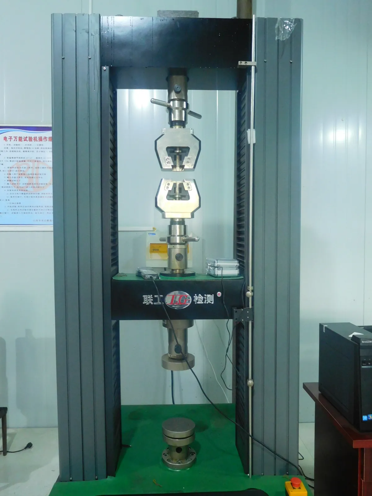 Floor-standing universal testing machine with tensile grips in a white-walled lab, a computer to the right