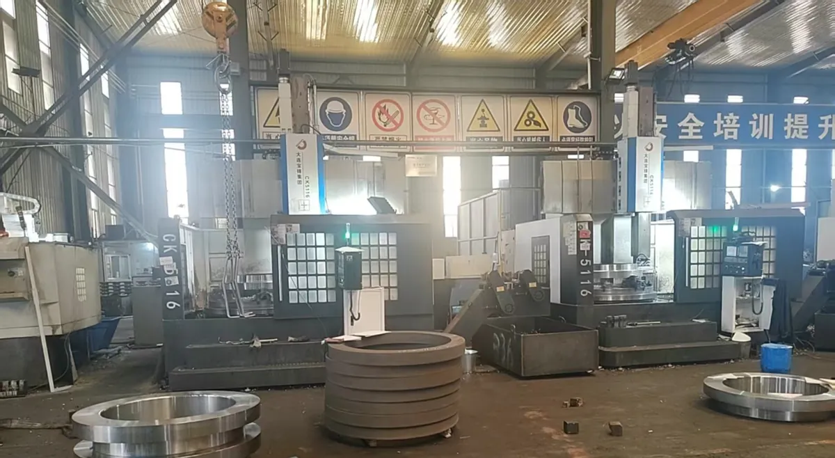 Two CNC vertical lathes under a row of safety signs, with machined rings and a stack of rough rings on the floor in front