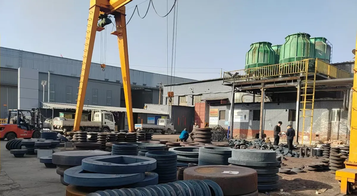 Yard of stacked forged ring and disc blanks under a yellow gantry crane, with green cooling towers on a platform