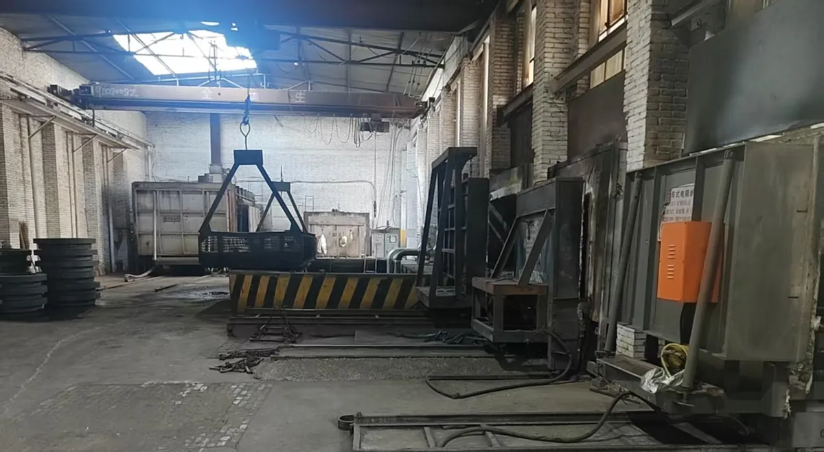Heat-treatment hall with furnace doors and a car-bottom furnace along the right wall, a loading basket on a crane
