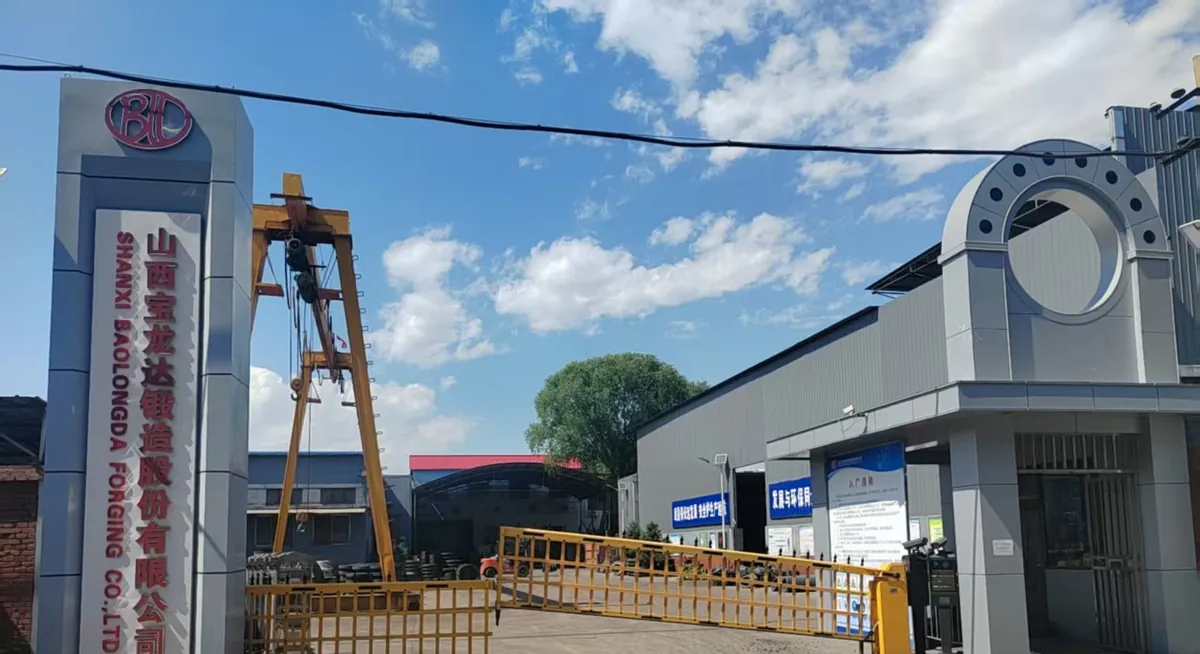 Factory gate with a tall grey pillar sign reading Shanxi Baolongda Forging Co., Ltd., a yellow barrier gate and a gantry crane