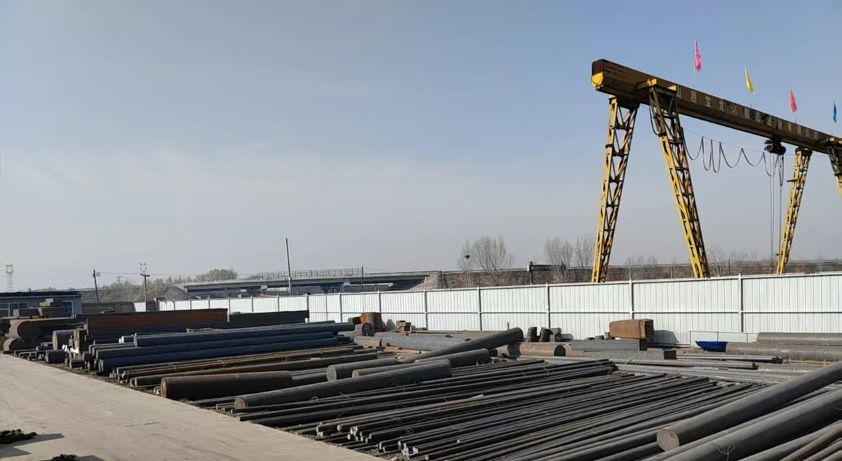 Outdoor yard of steel round bars and billets laid in rows under a yellow gantry crane whose beam carries the company name